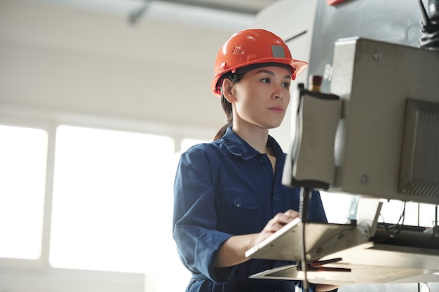 Grave giovane operatore asiatico in hardhat utilizzando il computer della macchina industriale durante la scelta delle configurazioni in officina