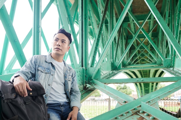 Serious young Asian traveler sitting with backpack under green bridge and looking around