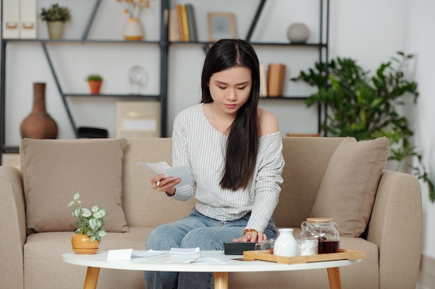 Serious young Asian woman checking utility bills or receipts, managing monthly expenses