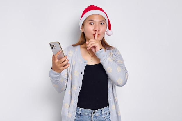 Serious young Asian woman in a Christmas hat holding mobile phone making silence gesture isolated on white background