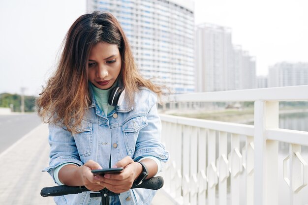 Serious young Asian woman standing on scooter and answering text messages from her friends