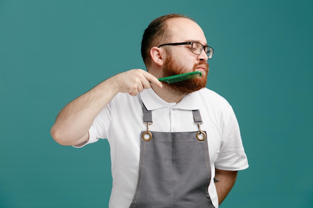 Serious young barber wearing uniform and glasses keeping hand behind back turning head to side combing his beard with teaser comb with closed eyes isolated on blue background