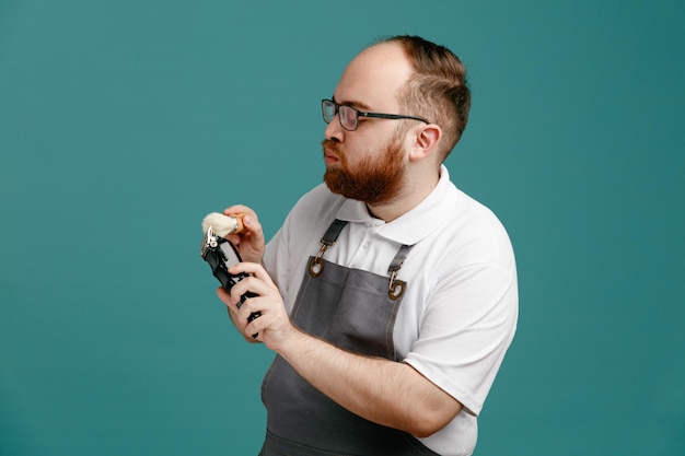 Serious young barber wearing uniform and glasses standing in profile view cleaning hair trimmer with shaving brush isolated on blue background