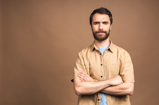 Serious young bearded man. Portrait of handsome young man in casual keeping arms crossed while standing against beige background.