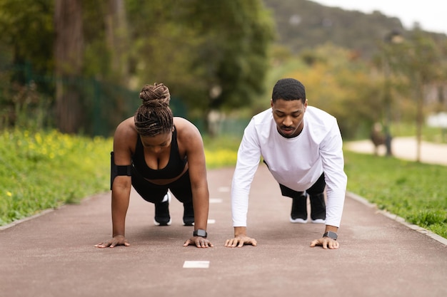 Serious young black couple do pushups and look at phone enjoy workout in summer park