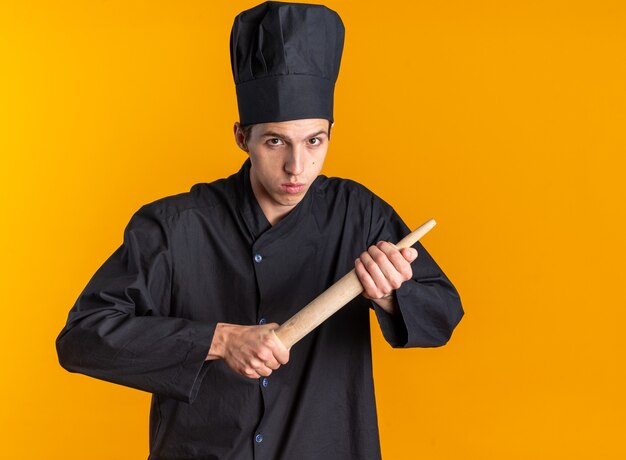 Grave giovane maschio biondo cuoco in uniforme da chef e berretto guardando la telecamera tenendo il mattarello con entrambe le mani isolate sulla parete arancione