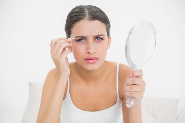 Serious young brown haired model in white pajamas plucking her eyebrows