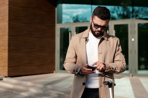 Serious young businessman in beige trenchcoat and sunglasses scrolling in smartphone while standing against entrance of modern office building