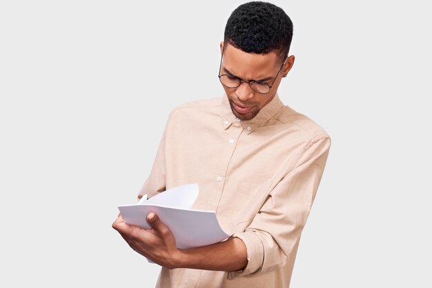 Serious young businessman in casual shirt analyzing financial reports posing on white studio background African American male student checking paper documents