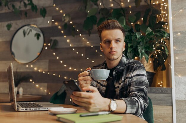 カフェでコーヒー ブレークを持ち、よそ見しながら携帯電話を閲覧している真面目な青年実業家