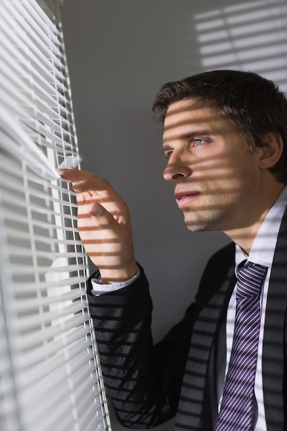 Serious young businessman peeking through blinds in office