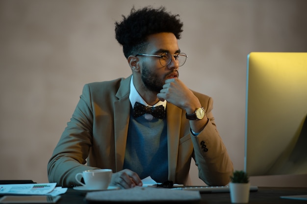 Serious young businessman sitting at the table in front of computer monitor and examining online work at office