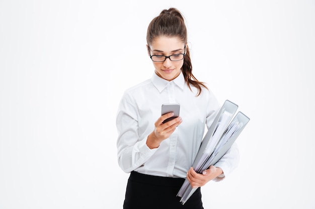 Serious young businesswoman in glasses holding folders and using smartphone over white wall