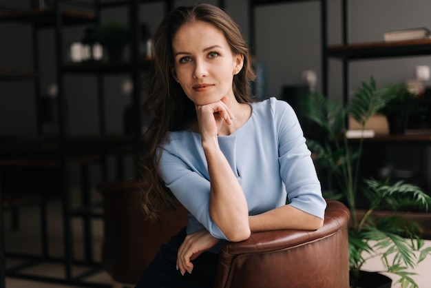 Serious young businesswoman wearing stylish light blue dress sitting at the chair in office