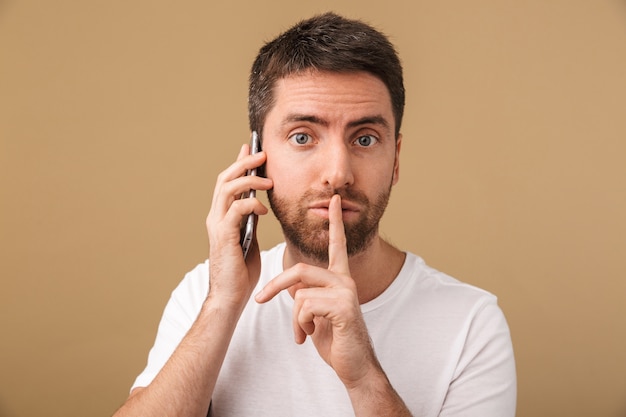 Photo serious young casual man talking on mobile phone isolated, showing silence gesture