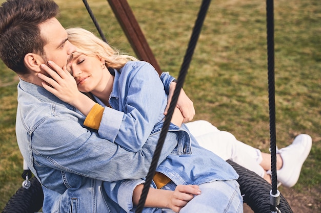 Serious young Caucasian man sitting with his beautiful sleepy serene blonde girlfriend on a swing