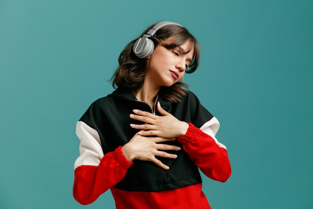 Serious young caucasian woman wearing headphones keeping hands on chest turning head to side looking down isolated on blue background