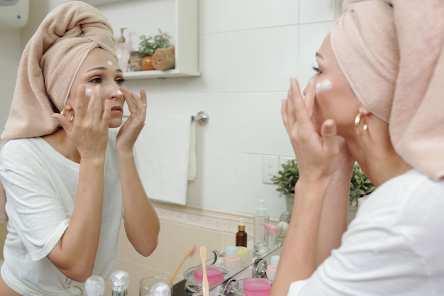 Photo serious young caucasian woman with hair wrapped in towel smoothing cream on face skin in front of mirror in bathroom