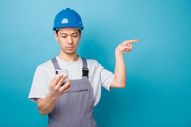 Serious young construction worker wearing safety helmet and uniform holding and looking at mobile phone pointing to side 