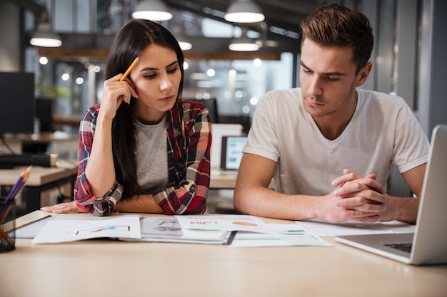 Serious young Coworkers sit by the table with documents and laptop in office