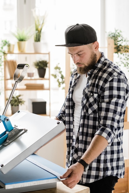 Serious young craftsman holding stripe with decor while printing it on new pet collar on special equipment by workplace