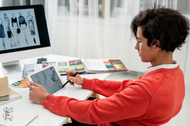 Serious young designer with pen pointing at fashion model on tablet screen while choosing new ones for seasonal collection