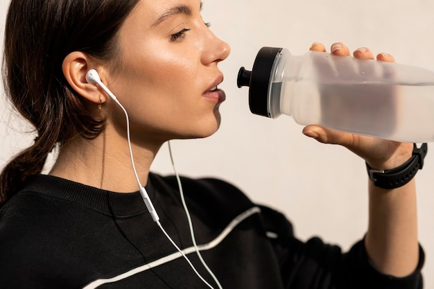 Serious young european woman athlete in sportswear and headphones drinking water from bottle relaxing