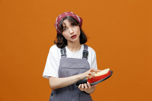 Serious young female cleaner wearing uniform and bandana cleaning tray with sponge looking at camera isolated on orange background