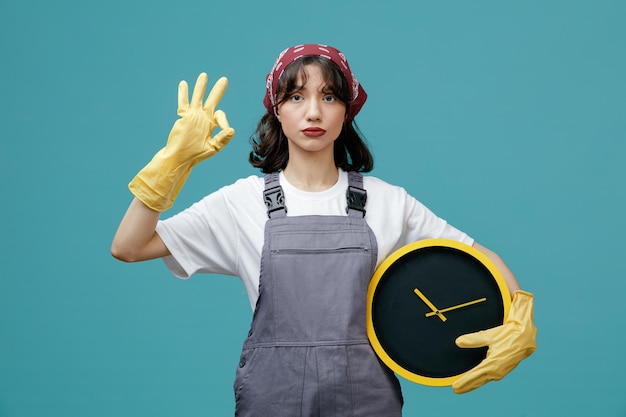 Serious young female cleaner wearing uniform bandana and rubber gloves holding clock looking at camera showing ok sign isolated on blue background