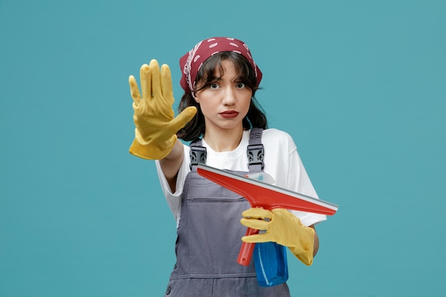 Foto seria giovane donna addetta alle pulizie che indossa bandana uniforme e guanti di gomma che tengono tergicristallo e detergente guardando la fotocamera che mostra il gesto di arresto isolato su sfondo blu