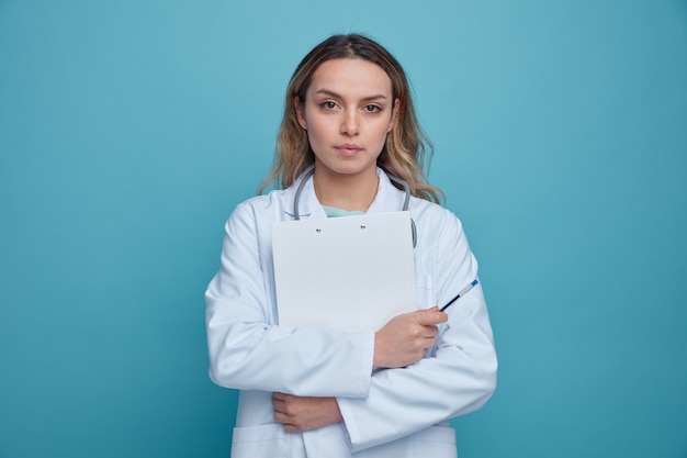 Serious young female doctor wearing medical robe and stethoscope around neck holding pen hugging clipboard 