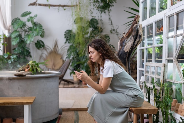 Serious young gardener woman owner holding smartphone reading media news sitting in home garden