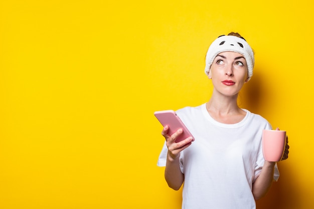 Serious young girl in a bandage looks to the side with a phone and a cup of coffee on a yellow background