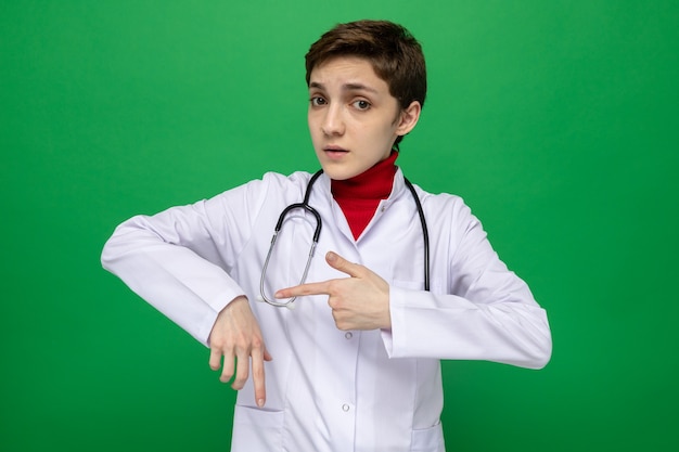 Serious young girl doctor in white coat with stethoscope around neck pointing at her hand reminding about time standing on green