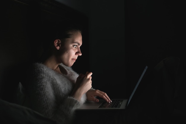 Serious young girl sitting in the dark at night on the bed and watching a movie on the laptop