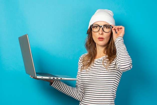 Serious young girl in a white hat with a laptop holding on to glasses on blue