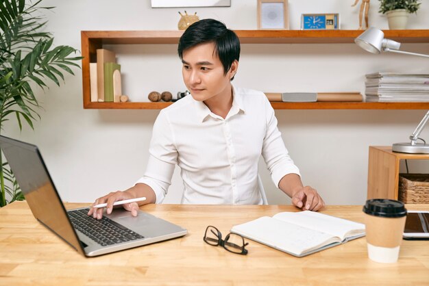 Serious young handsome businessman checking information on laptop screen and taking notes in planner