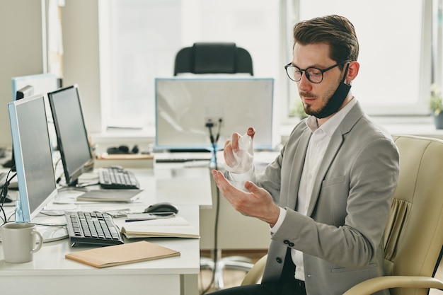 手に消毒剤をスプレーあごにマスクを持つ深刻な若いハンサムな男