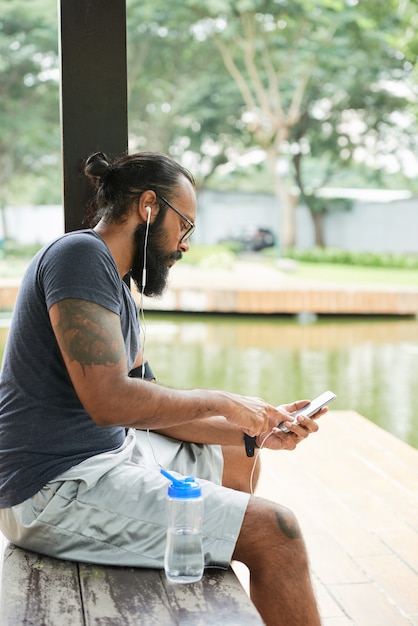 Serious young Indian bearded sportsman with tattoos sitting on bench at pond and listenign to music via phone app