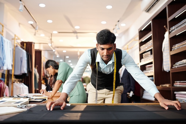 Serious young Indian tailor masuring black wool fabric to make bespoke suit for client