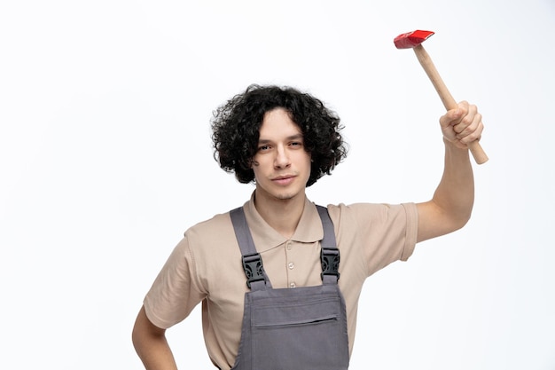 Serious young male construction worker wearing uniform looking at camera raising hammer up isolated on white background