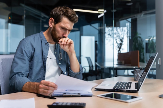 A serious young man accountant financier analyst auditor sits in the office at the table