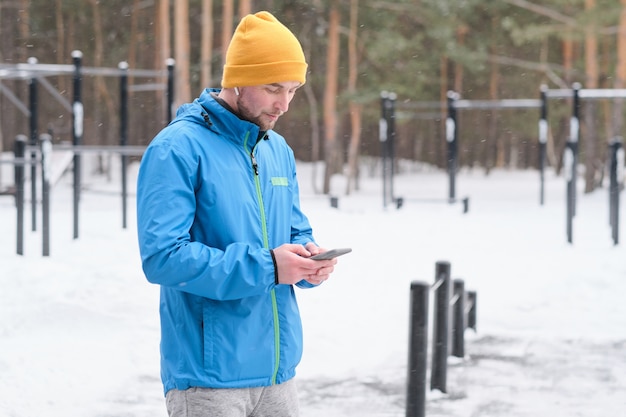 Серьезный молодой человек в яркой шляпе стоит на зимней спортивной площадке и с помощью смартфона делает заметки о тренировках