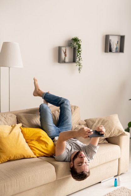 Serious young man in casual clothing sitting upside down on sofa and playing gadget game during home isolation