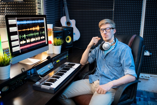 Photo serious young man in casualwear sitting in armchair by desk with computer monitor and keyboard while mixing sounds in studio