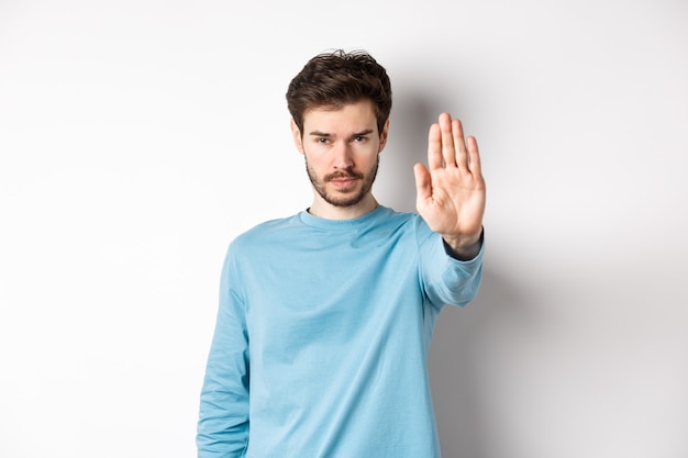 Giovane serio che sembra fiducioso con la mano tesa, dicendo stop, proibire qualcosa di brutto o dare avvertimento, in piedi su sfondo bianco