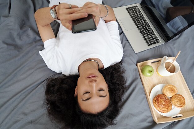 Serious young man lying on bed with tray of breakfast and ope laptop and texting friends