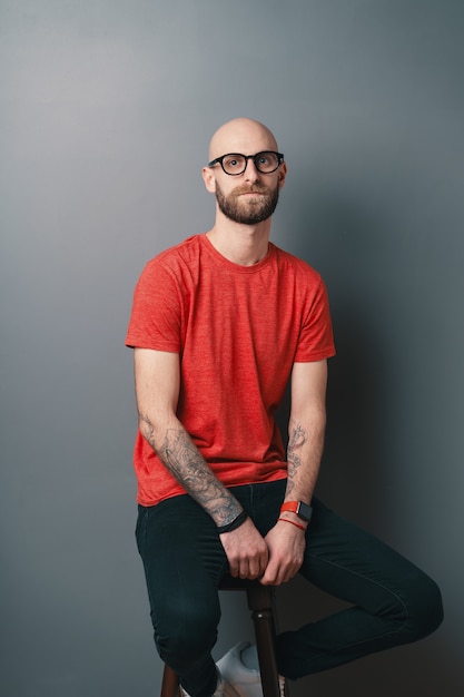 Serious young man in red t-shirt posing in the studio on gray background while sitting on a tall chair