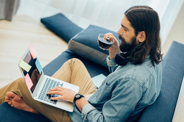 Photo serious young man with a cup of coffee sitting on the sofa with a modern laptop. sticky notes on the screen