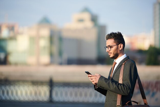 通りを歩きながら電話メッセージをチェックするスーツを着た真剣な若い中東のビジネスマン